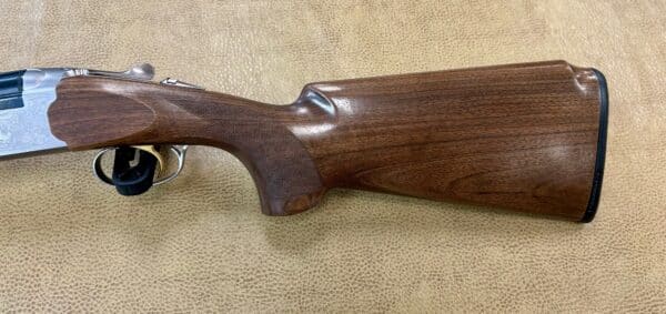 Side view of a wooden rifle stock with metal details on a textured beige background.