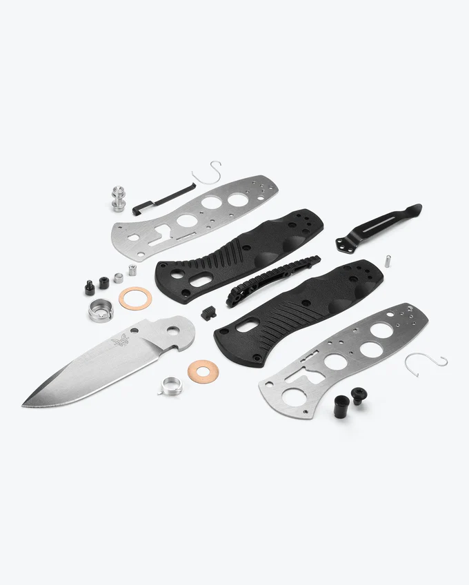 Disassembled knife components arranged on a light surface, including a blade, handle scales, screws, and hardware pieces.