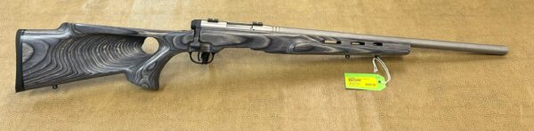 A bolt-action rifle with a gray laminated stock rests on a textured surface with a price tag.