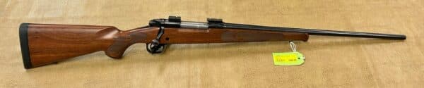 The image shows a horizontal view of a wooden bolt-action rifle against a textured brown background. The rifle has a lustrous dark wood stock with a pronounced grip and a smooth finish. The barrel, which is long and slender, displays a shiny black metal finish. The scope mounts are visible on top of the rifle, and a yellow price tag is attached, indicating the item's price. The lighting highlights the wood grain and reflects off the metal components, creating a visually rich appearance.