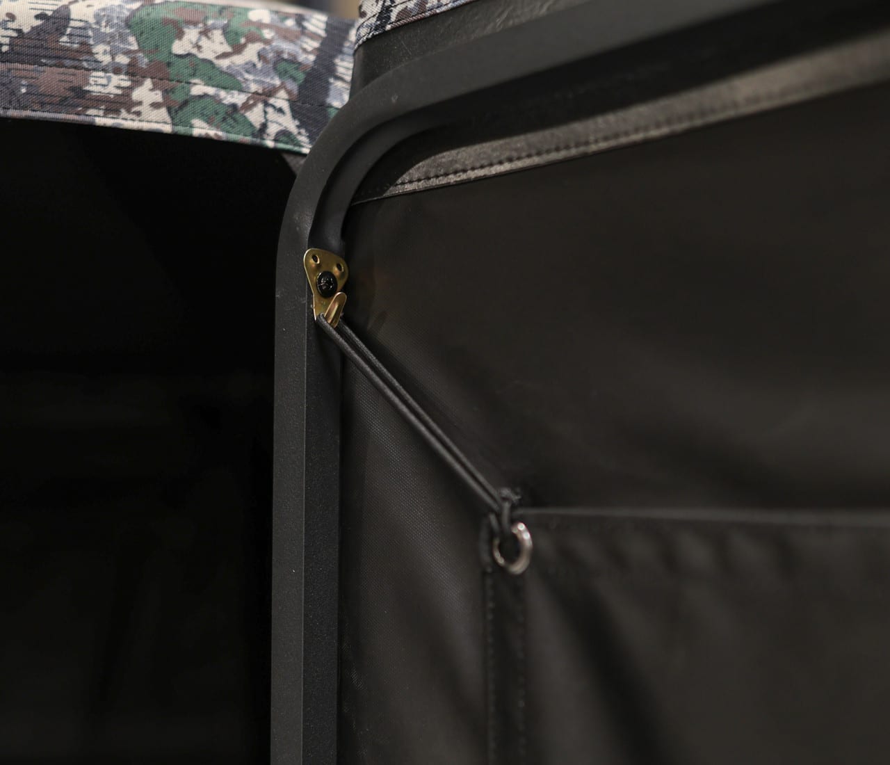 Close-up of the interior of a hunting blind, showing a black fabric wall with a bungee cord attached to a metal ring and a small metal clasp, with camouflage material visible in the upper left.