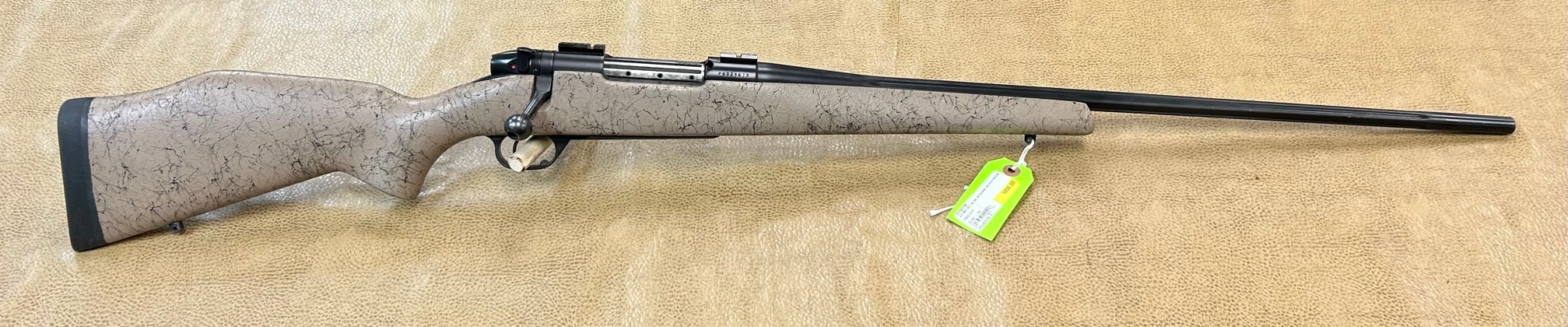 Side view of a bolt-action rifle with a textured gray stock and a long black barrel, resting on a light-colored surface.